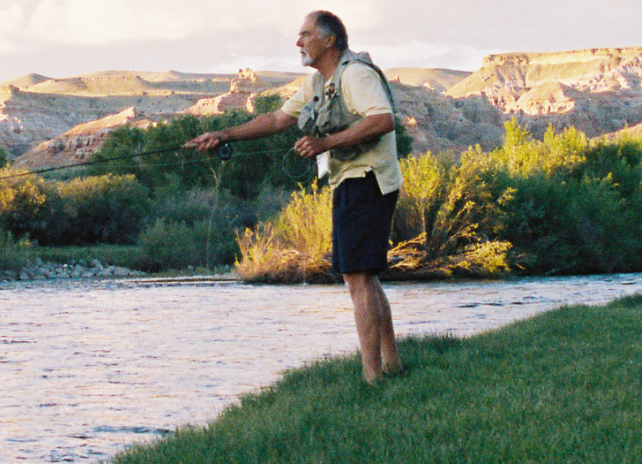 A man fishes the Green River along a grassy stretch of shoreline with mountains in the background