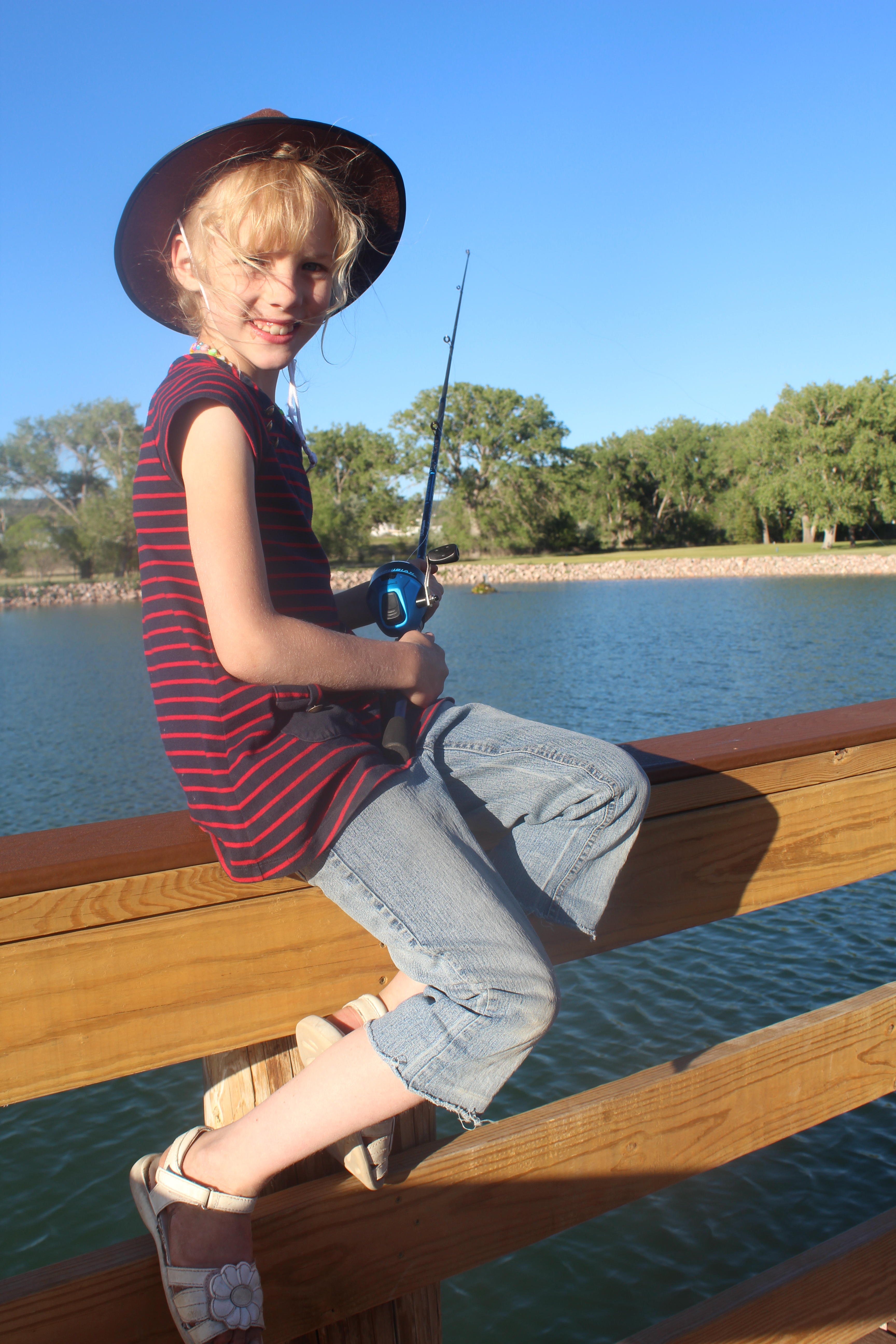 A young girl sits on the railing by the pond with a spin fishing rod.