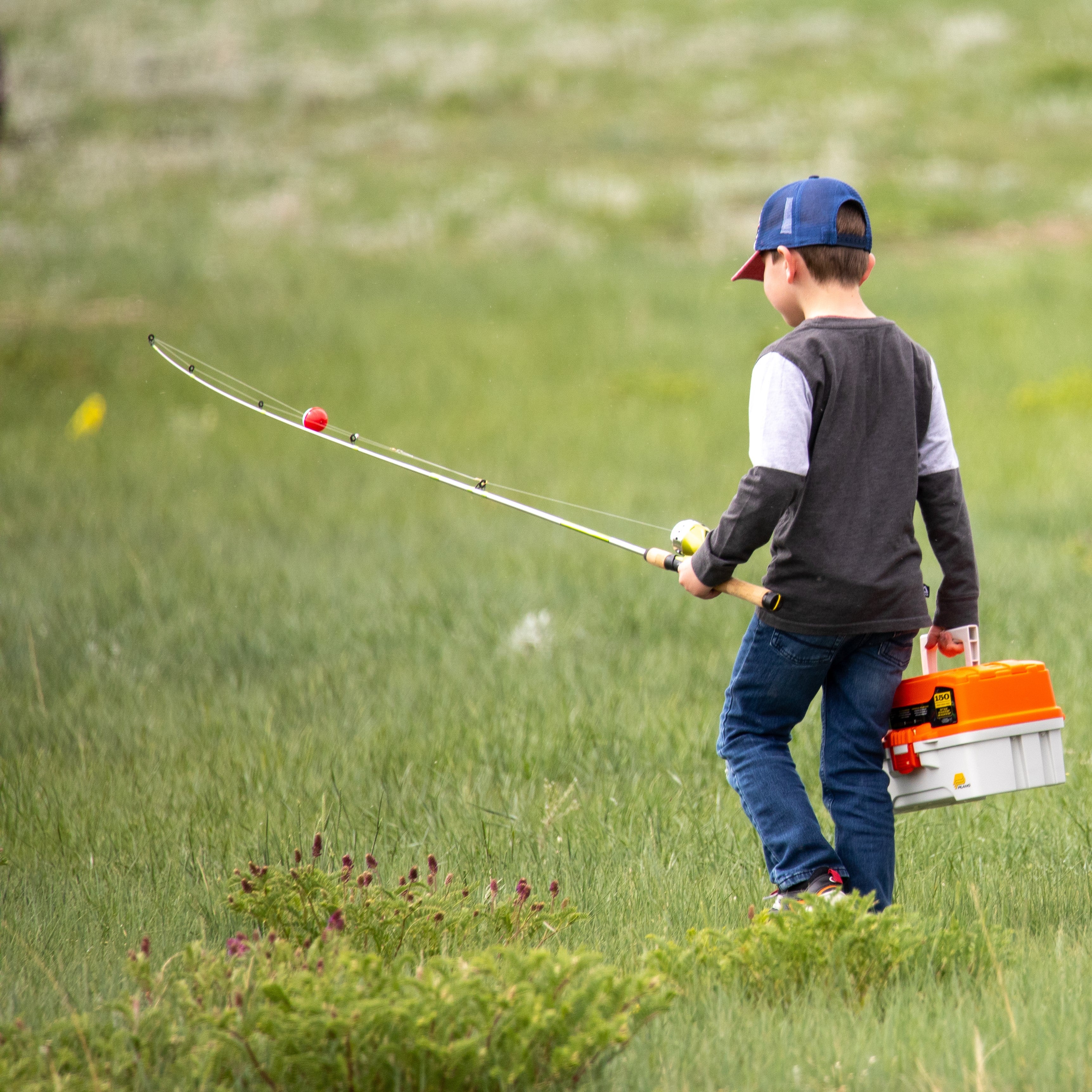 A kid with a fishing pole and tacklebox walks through a field