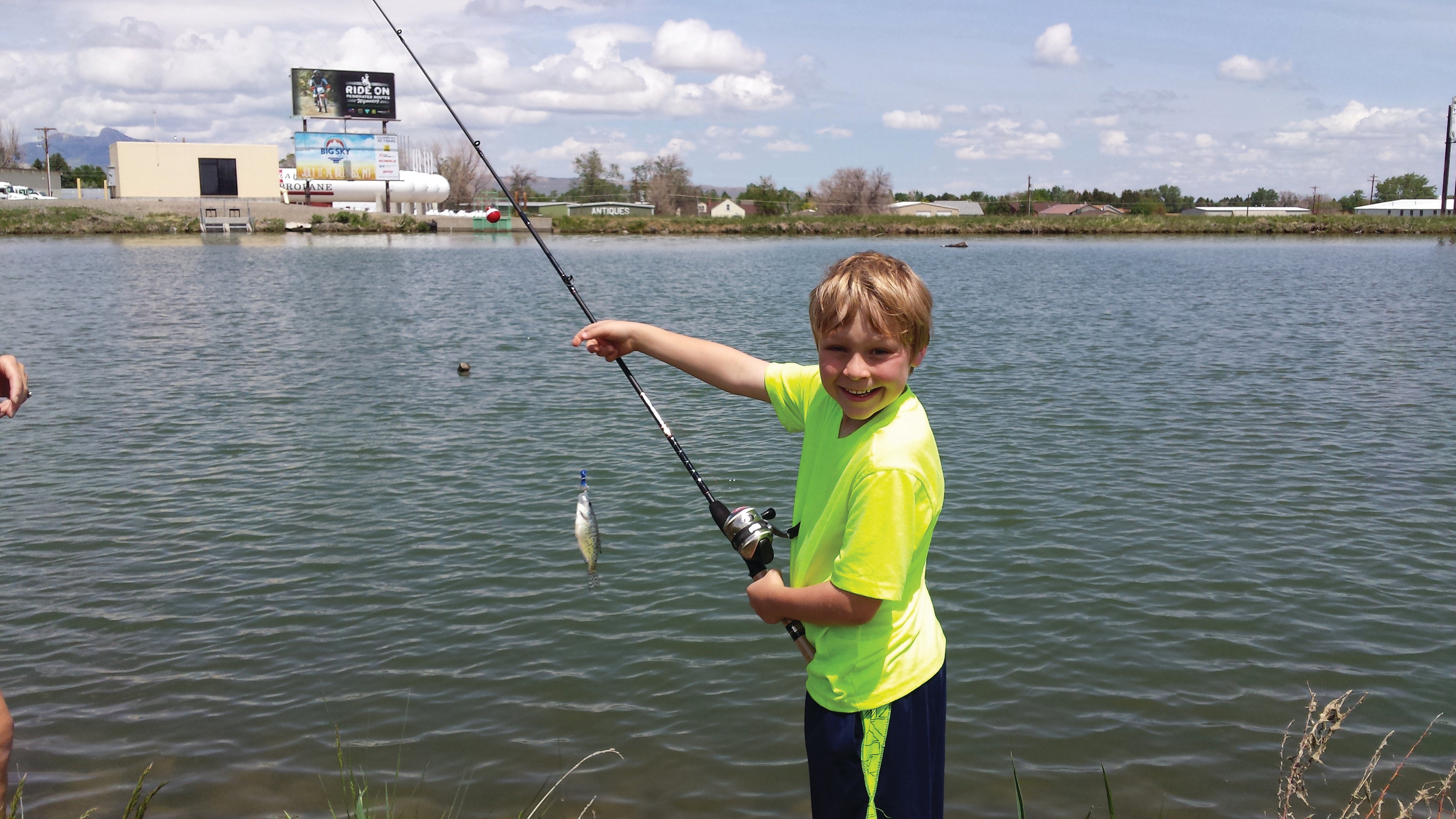 A young angler in a bright green shirt with a fishing rod shows off a fish on the end of his line.