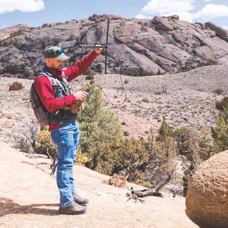 Lucas Olson, Game and Fish quantitative wildlife biologist, stands on a rocky landscape and uses radiotelemetry to locate a mule deer GPS collar in the Sweetwater Rocks area.