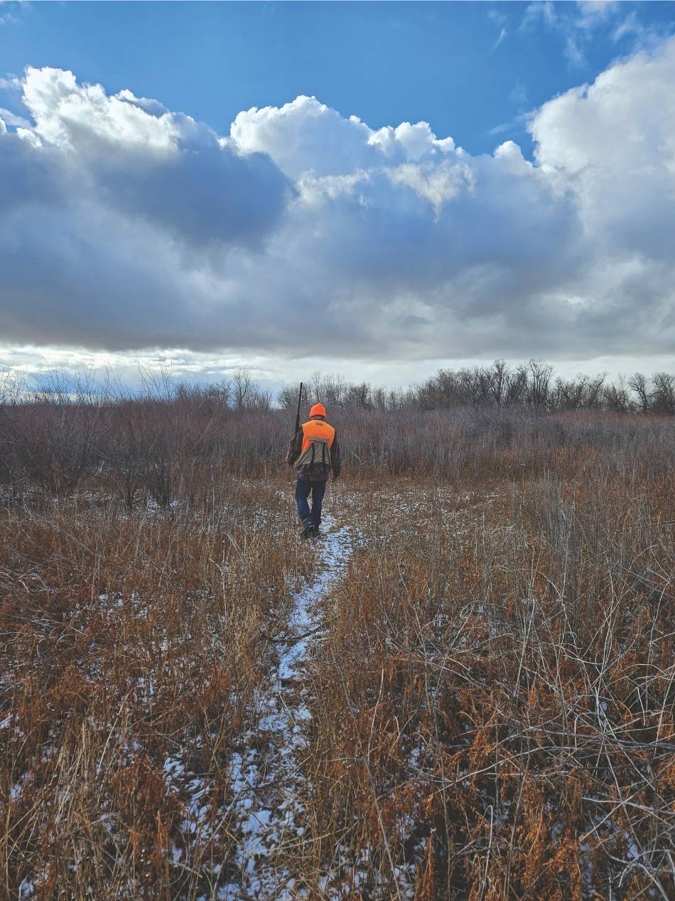 A hunter walks down a snowy path with a cloudy sky in the distance.