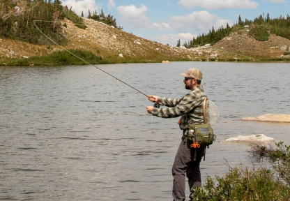 Man in long sleeve plaid shirt and hat casts while fly fishing from shore at an alpine lake. 