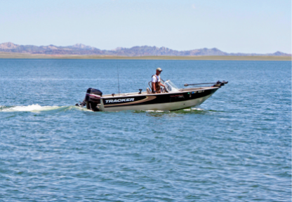 Man in overalls drives a motorized fishing boat on a lake on a clear day with mountains in the distance.