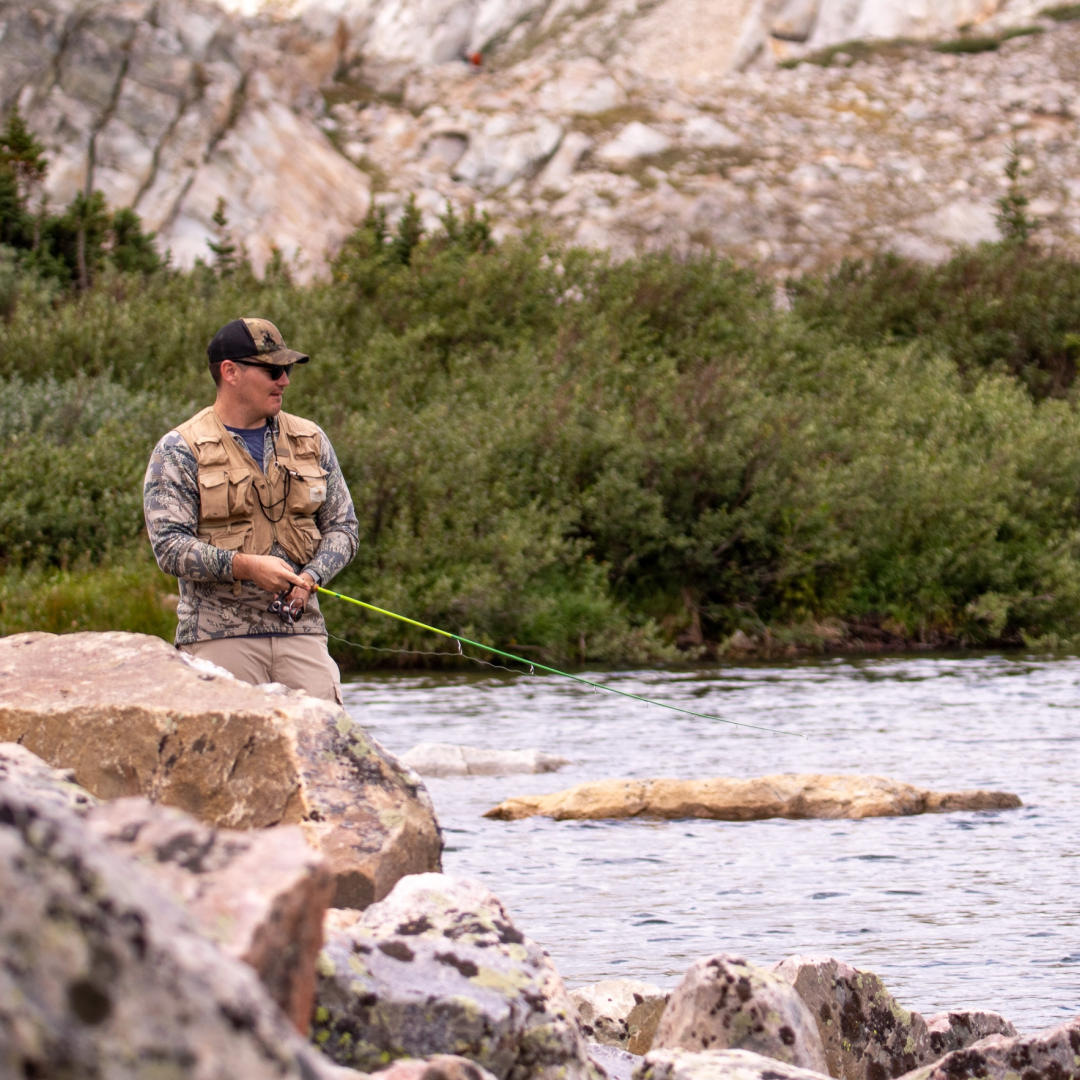 Male angler in a fishing vest, hat, and camo long-sleeve shirt spin fishes along a Wyoming stream