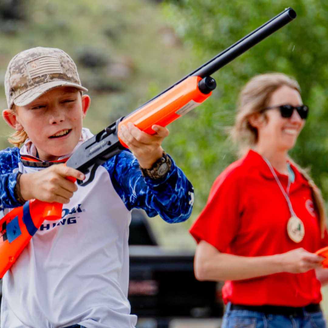 A hunter education student practices safe zones of fire with an inert shotgun while a hunter education instructor coache
