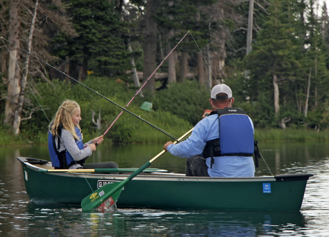 A man and girl spin fish from a green canoe on a lake bordered by evergreen trees