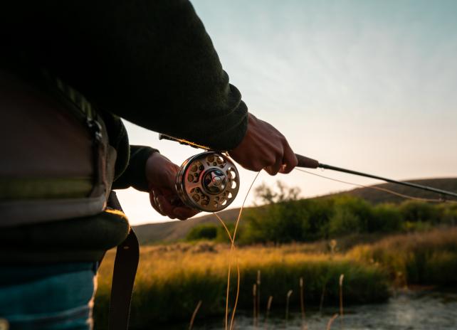 Close up of a female angler holding a fly fishing rod and reel on the edge of a creek, backlit in early morning light