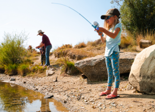 A youth angler reels in a fish on a summer day at CCC ponds in Pinedale, Wyoming while her mother assists her brother with fishing in the background