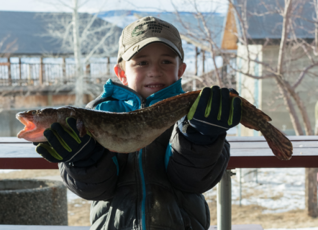 A youth angler in a winter jacket, gloves, and a baseball hat holds a burbot for a photo at the Burbot Bash Fishing Derby.