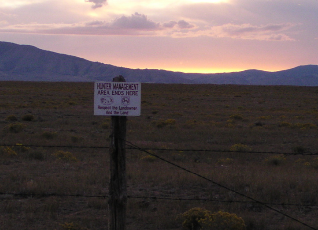 A Hunter Management Area sign posted on a barbed wire fence in front of a mountainous landscape during sunset.