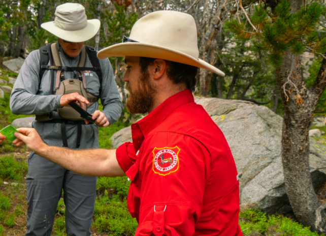 A game warden checks a fishing license of two male anglers hiking to fish backcountry lakes