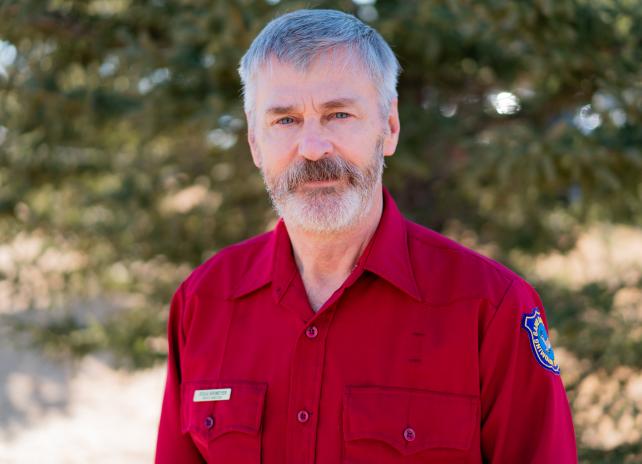 Doug Brimeyer A portrait photo of Deputy Director of External Affairs Doug Brimeyer in Wyoming Game and Fish Department red-shirt uniform with bokeh of evergreen trees in the background