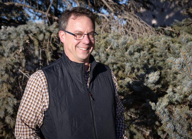 A portrait of Jeff Davis, Deputy Director of External Affairs standing in front of a wall of evergreen branches in the background.