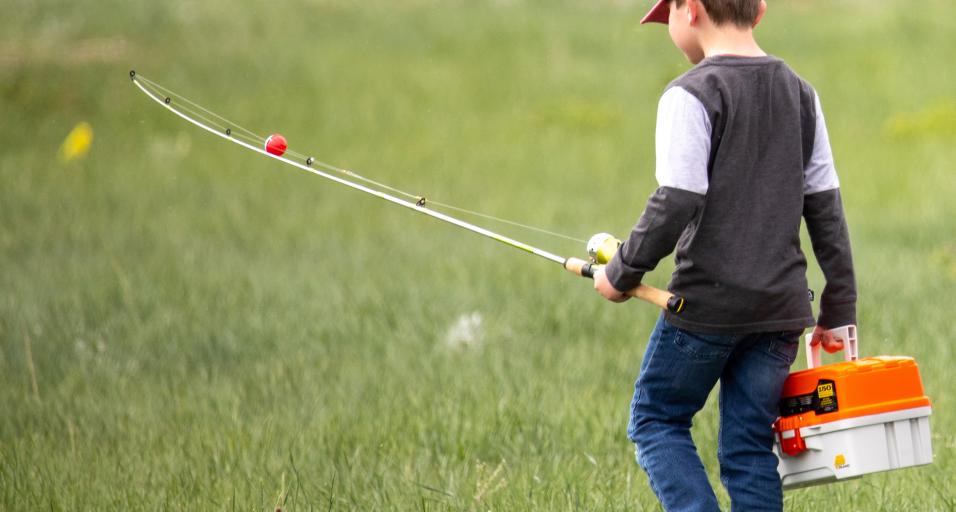 A kid with a fishing pole and tacklebox walks through a field