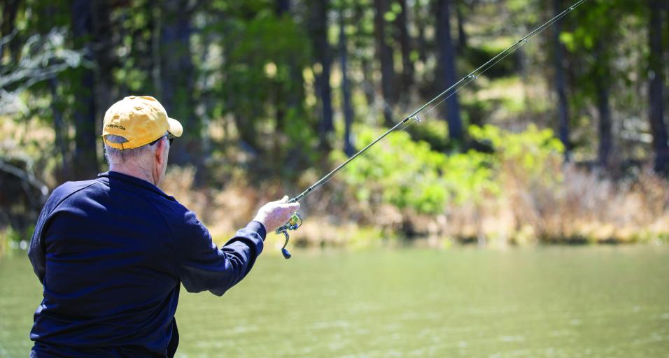 A male angler with a blue shirt and yellow hat fishes with a spin casting rod from shore.