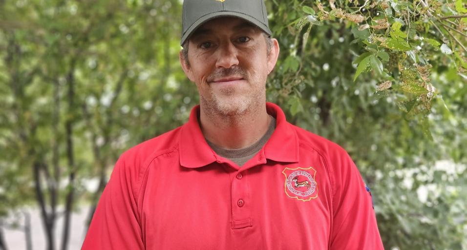 Wyoming Game Warden Brady Frude stands in front of trees with his red uniform shirt, green hat and a smile