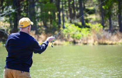 A male angler with a blue shirt and yellow hat fishes with a spin casting rod from shore.