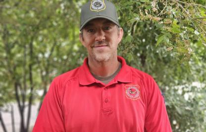 Wyoming Game Warden Brady Frude stands in front of trees with his red uniform shirt, green hat and a smile