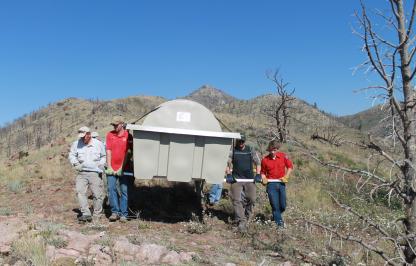 Four people carrying a large gray container through a hilly area with dry brush and a couple dead trees.