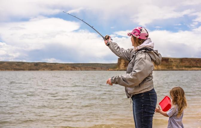 A mother casts a fishing pole for her daughter as the daughter holds a red bucket at Flaming Gorge Reservoir