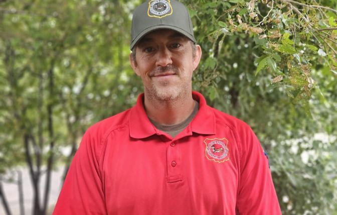 Wyoming Game Warden Brady Frude stands in front of trees with his red uniform shirt, green hat and a smile