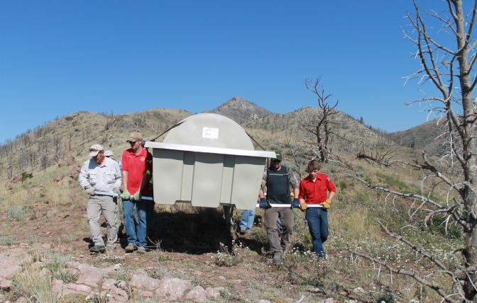 Four people carrying a large gray container through a hilly area with dry brush and a couple dead trees.